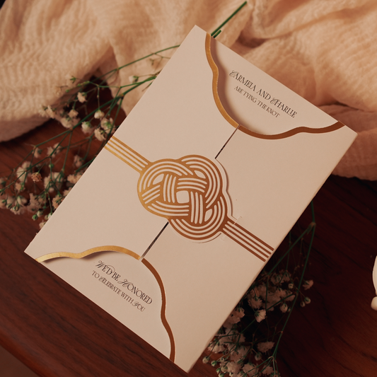 White gate-fold invitation with gold foil knot accent laying on top of a bed of babys breath flowers